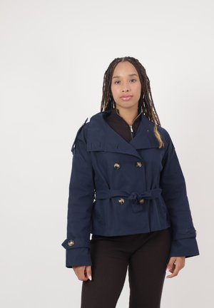 Femme aux longues tresses portant une veste bleu marine croisée avec ceinture et un pantalon noir, debout devant un fond blanc uni.