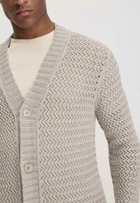 Helle graue Strickjacke mit Fischgrätenmuster, V-Ausschnitt und zwei Knöpfen vorne. Strukturiertes Material mit gerippten Kanten.