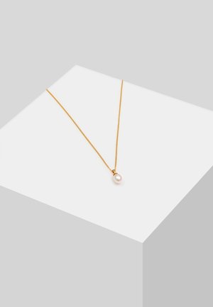 Collier délicat en chaîne d’or avec un pendentif unique en perle blanche présenté sur un piédestal géométrique blanc sur fond gris.