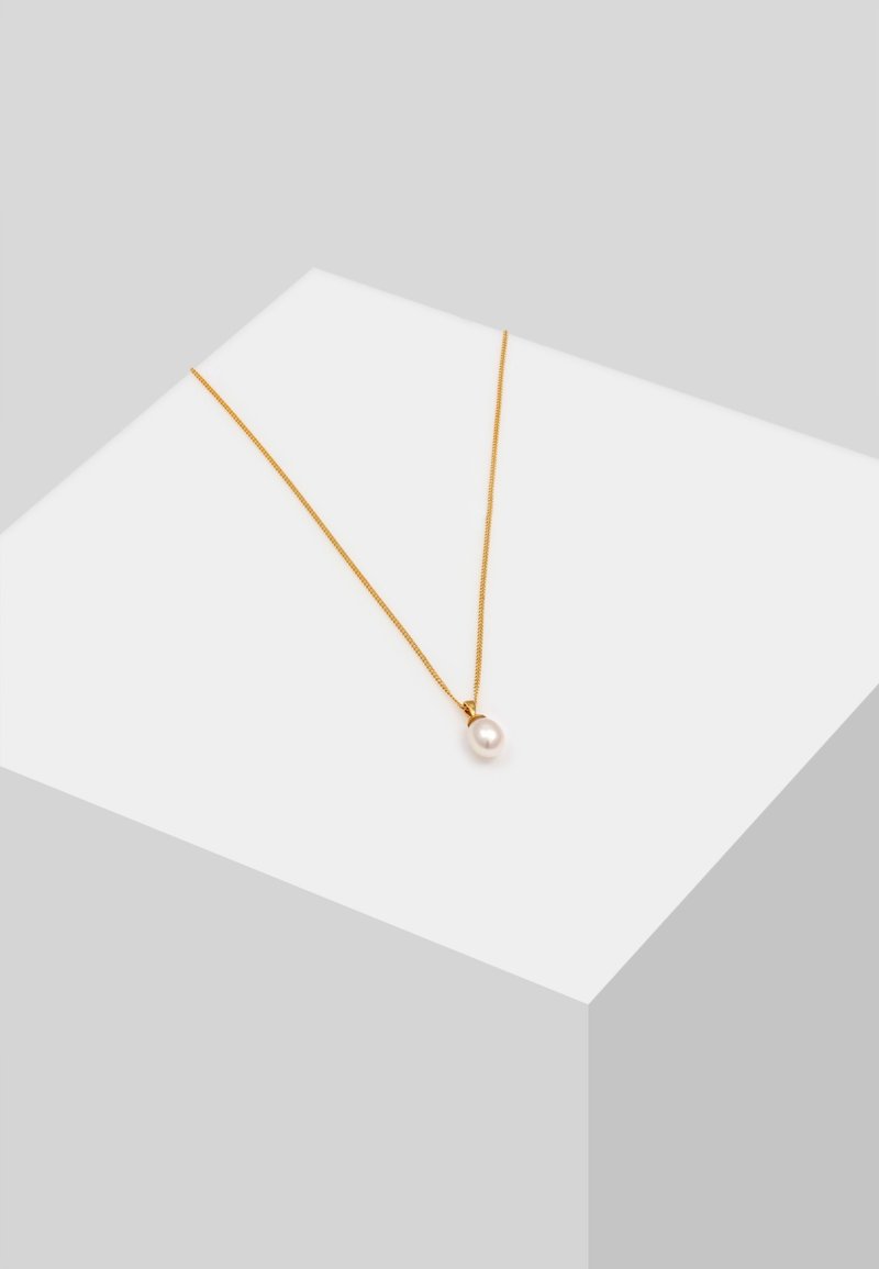 Collier délicat en chaîne d’or avec un pendentif unique en perle blanche présenté sur un piédestal géométrique blanc sur fond gris.