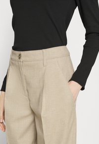 Pantaloni beige a spina di pesce con vita alta, caratterizzati da una chiusura con bottone e tasche laterali, abbinati a un top nero a coste attillato.