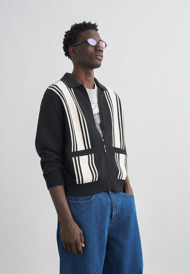 Νεαρός άνδρας με στρογγυλά γυαλιά, μαύρο πουλόβερ με φερμουάρ και λευκές κάθετες ρίγες, και μπλε τζην, στέκεται μπροστά σε απλό φωτεινό φόντο.