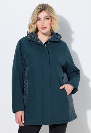 Lyshåret kvinde iført en mørkteal hooded vinterjakke med quiltede sidepaneler, stående med den ene hånd i lommen mod en ensfarvet lys baggrund.