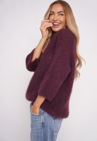 Kobieta z długimi blond włosami, ubrana w fioletowy, puszysty sweter i niebieskie dżinsy, uśmiecha się, trzymając jedną rękę przy twarzy, a drugą w kieszeni.