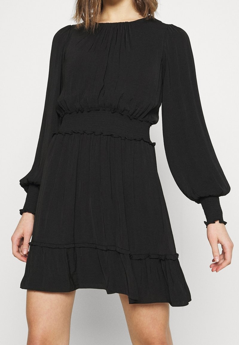 Femme portant une robe noire à manches longues avec taille et poignets élastiques, jupe plissée et détails à volants à l’ourlet et à la taille.