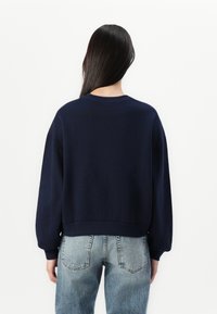 Marineblauwe sweatshirt met een ronde hals, verlaagde schouders en ribgebreide manchetten. Draagt bij lichte denim jeans met achterzakken.
