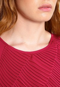 Bordó blůzka s texturovanými sklady na přední straně a kulatým výstřihem. Látka vypadá hladce s mírným leskem, což zdůrazňuje detailní švy.