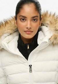 Hvid dunjakke med hætte foret med pels, quiltet tekstur og en sort lynlås-sweatshirt under. Har en forlynlås og snøretræk.