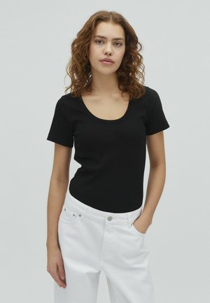 Tânără cu păr creț castaniu, purtând un top negru cu mâneci scurte și pantaloni albi, stând cu o mână în buzunar pe un fundal gri.