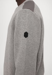 Pulover tricotat gri cu decolteu rotund, textură canelată și reparații gri pe umeri. Logo hexagonal negru pe mâneca stângă.