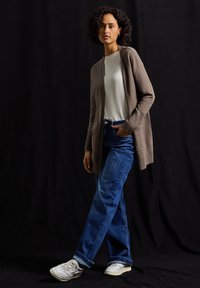 Jasnobrązowy dzianinowy kardigan z otwartym przodem, zestawiony z niebieskimi dżinsami, białą koszulką i białymi trampkami. Prosty, luźny krój i teksturowany materiał.