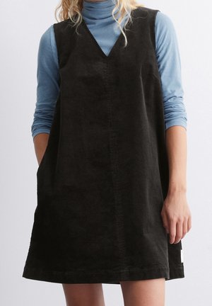 Mujer con vestido de pana negro sin mangas sobre una camisa de cuello alto azul claro de manga larga, de pie frente a un fondo liso.
