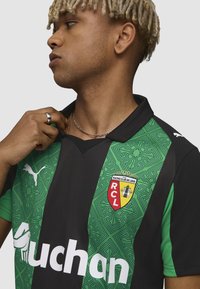 Mladý človek s svetlými kučeravými vlasmi má na sebe zeleno-čierny futbalový dres s emblémom Racing Club de Lens a logom sponzora.