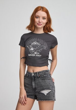 Cropped top grigio scuro con maniche corte, caratterizzato da una stampa grafica bianca, abbinato a shorts in denim neri strappati a vita alta.