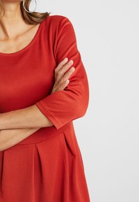 Punainen, pitkähihainen mekko, jossa on pyöreä kaula-aukko, valmistettu pehmeästä kankaasta. Ominaisuuksiin kuuluu pleatattu vyötärö ja sileä pinta.