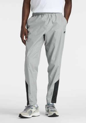 Mann trägt hellgraue Sporthose mit schwarzen Einsätzen an den Waden und graue New Balance-Sneaker, steht mit Händen in den Taschen.