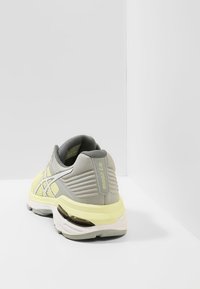 Chaussure de sport grise avec des accents jaunes, tige en maille texturée, semelle rembourrée et détails réfléchissants à l'arrière. Vue arrière affichée.