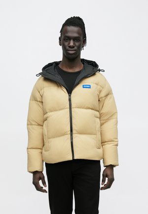 Μπεζ μπουφάν τύπου puffer με μαύρη κουκούλα, φερμουάρ μπροστά και μπλε λογότυπο. Διαθέτει καπιτονέ σχέδιο και λεπτή εφαρμογή, φορεμένο πάνω από μαύρο πουκάμισο.