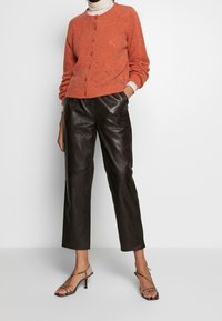 Cardigan en tricot orange rouille avec boutons, associé à un pantalon court en cuir noir taille haute et des sandales à talons à lanières.