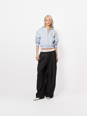 Blondynka stoi, ubrana w jasnoniebieską krótka bluzę na suwak, szerokie czarne spodnie, białe spiczaste buty i biały pas widoczny nad spodniami.