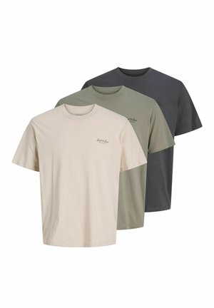 Jeune homme debout regardant sur le côté, portant un t-shirt beige à manches courtes, un jean noir ample et des chaussures marron sur un fond clair uni.