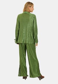 Conjunto de satén texturizado verde que presenta una blusa de manga larga con puños atados y pantalones de pierna ancha con aberturas laterales; acabado suave y brillante.