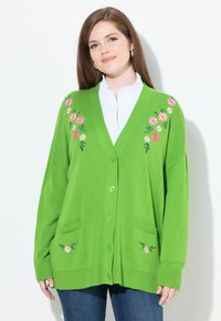 Kvinna som bär en klargrön kofta med rosa och vita blommönster över en vit skjorta och blå jeans mot en vit bakgrund.