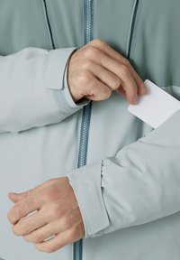 Lichtblauwe jas met een rits, voorzien van een zak op de mouw voor een pasje. Glad materiaal en verstelbare manchetten.
