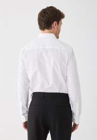 Férfi fehér hosszú ujjú ingben, fekete öltönynadrágba tűrve, háttal áll egy sima fehér háttér előtt.