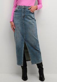 Falda maxi de mezclilla en azul claro, con una abertura frontal, cintura alta y diseño de cinco bolsillos. Combinada con botas negras hasta la rodilla.