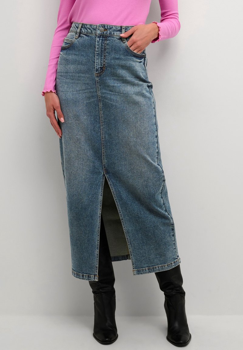Falda maxi de mezclilla en azul claro, con una abertura frontal, cintura alta y diseño de cinco bolsillos. Combinada con botas negras hasta la rodilla.