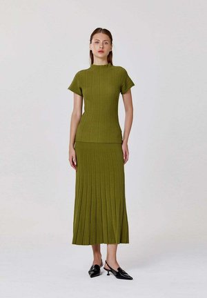 Femme debout portant un haut côtelé à manches courtes vert olive et une jupe midi plissée assortie avec des chaussures slingback noires à petit talon.