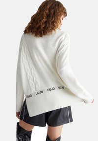 Witte gebreide trui met een kabelbrei detaillering aan de ene kant en een zwarte band met merknaam, gepresenteerd met bijpassende zwarte shorts.