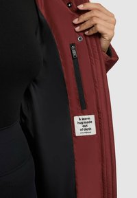 Maroon jacket made of nylon with a black interior, featuring a side zip pocket and a label stating "A warm hug made out of cloth."