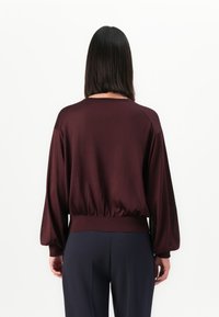 Persoon met steil zwart haar draagt een losse, lange mouw bordeauxrode top en donkere broek, staand tegen een vlakke witte achtergrond.