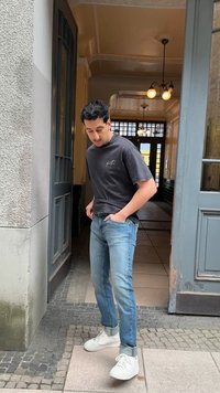 Άνδρας με μαύρο μπλουζάκι, μπλε τζιν και λευκά αθλητικά παπούτσια στέκεται στην πόρτα με το ένα χέρι στην τσέπη, κοιτάζοντας προς τα κάτω.