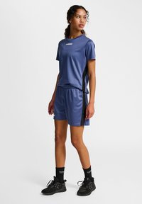 Blauwe sportset bestaande uit een kortzamige, vochtafvoerende top en shorts met elastische tailleband en zwarte zijpanelen, aangevuld met zwarte sneakers.
