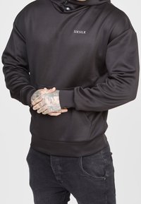Sudadera negra de tela suave con un bolsillo frontal, capucha ajustable con botones a presión y un pequeño logo en el pecho.