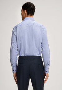 Camicia azzurra a maniche lunghe con colletto a punta, polsini con bottone e tessuto testurizzato, abbinata a pantaloni dritti scuri.