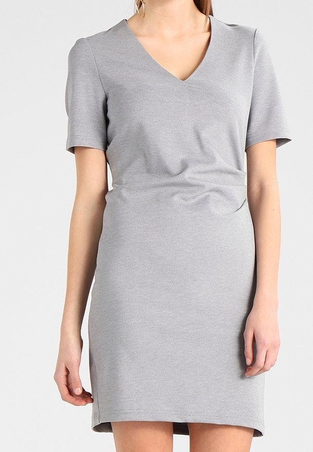 Femme portant une robe simple gris clair, manches courtes, longueur genou, avec un décolleté en V, sur un fond uni.