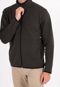 Giacca con zip verde oliva scuro in materiale morbido, con colletto alto, tasche laterali e dettagli a contrasto neri.