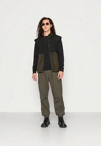 Vest i svart og olivengrønt med hette, kombinert med mørke cargobukser. Glatt stoff med glidelåsdetaljer og lommer. Svarte kampstøvler.