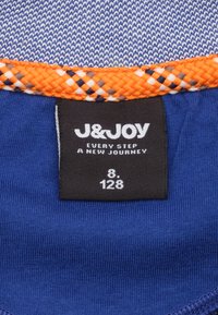 Étiquette de vêtement bleue avec un bord tissé orange et noir. L'étiquette affiche "J&JOY," taille "8," et numéro de style "128."