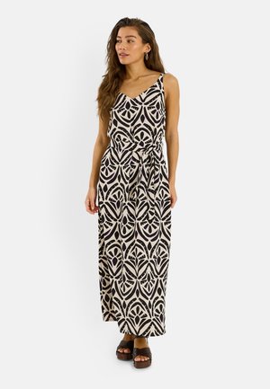 Mujer con cabello largo que lleva un vestido largo sin mangas de patrón negro y beige con cinturón en la cintura y sandalias oscuras con plataforma, de pie sobre fondo blanco.