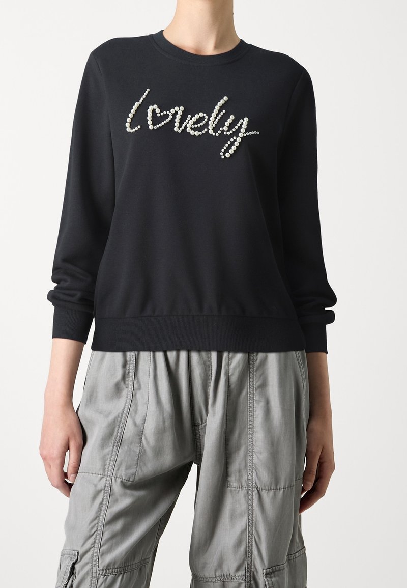 Sweat-shirt noir à col rond et manches longues, orné de perles formant le mot « lovely » sur le devant. Pantalon cargo gris.