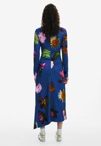 Langärmliges Kleid mit floralem Druck auf blauem Stoff, passgenauer Form, gerafften Details am Rücken und knielanger Länge.