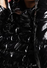 Černá péřová bunda z lesklého materiálu s prošívaným vzorem a stahující texturou. Detailní záběr zdůrazňuje šití a lesk.