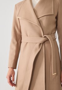 Cappotto avvolgente beige in tessuto morbido, con colletto ampio, maniche lunghe, vita con cintura e un accento dorato sul lato sinistro.