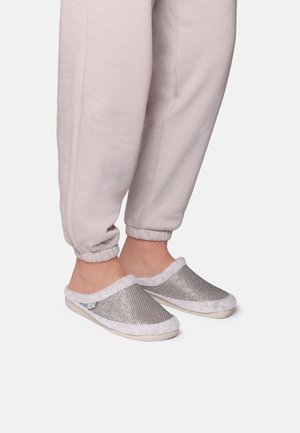 Silver vävda slip-on tofflor med vit lurvig foder och beige sula, matchade med ljusgrå joggerbyxor.
