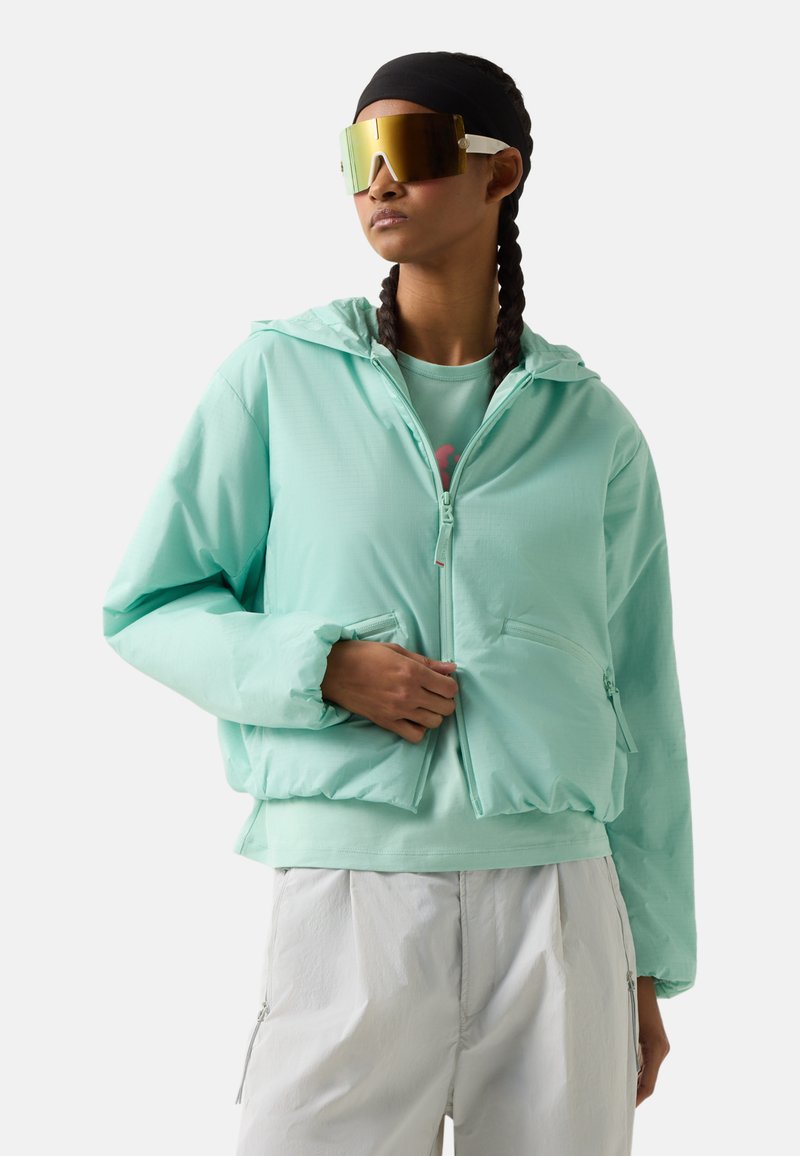Woman with braided hair wears mint green hooded jacket, matching shirt, white pants, and large reflective sunglasses, posing against white background.